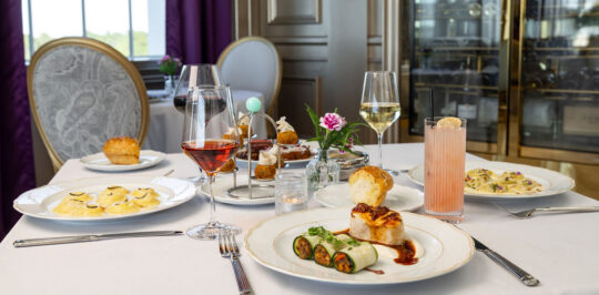 A table set with an assortment of plated gourmet dishes, desserts, and drinks in an elegant dining room with purple curtains and classic decor.