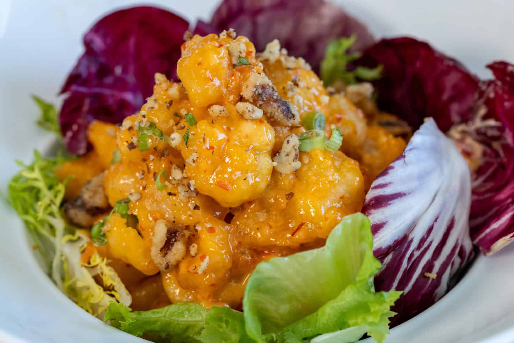 A close-up of a salad with greens and purple radicchio, topped with battered, sauced shrimp and sprinkled with chopped nuts and herbs.