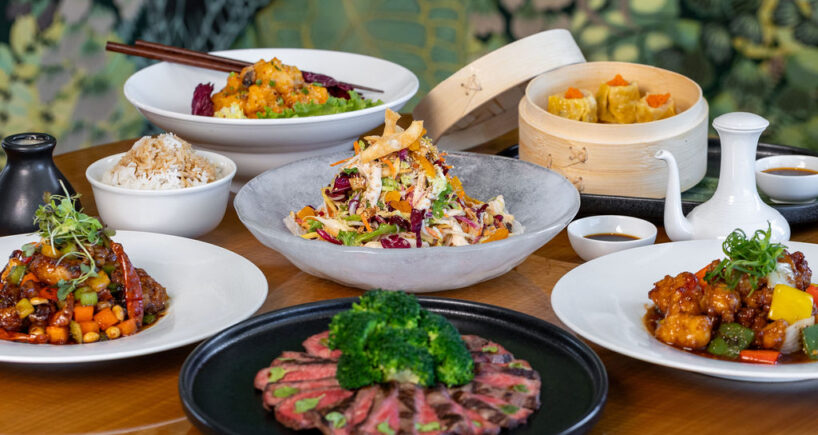 A selection of Asian dishes on a table, including sliced beef with broccoli, mixed salads, stir-fried vegetables, steamed dumplings, rice, and various sauces.