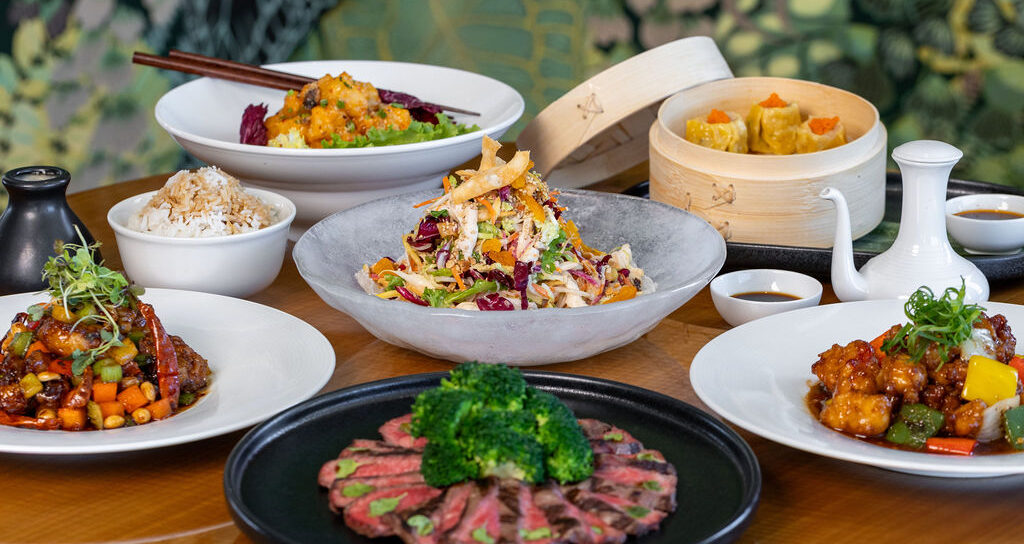A selection of Asian dishes on a table, including sliced beef with broccoli, mixed salads, stir-fried vegetables, steamed dumplings, rice, and various sauces.