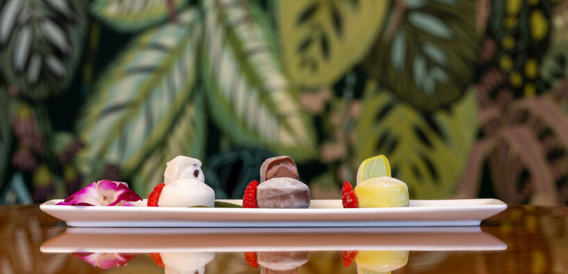 A rectangular white plate holds three assorted mochi desserts, garnished with berries and a flower, set against a leafy patterned background.