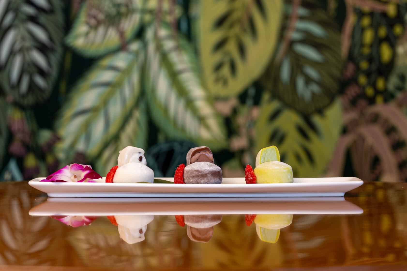 A rectangular white plate with four small, colorful desserts and garnishes, set on a reflective surface with a leafy patterned background.