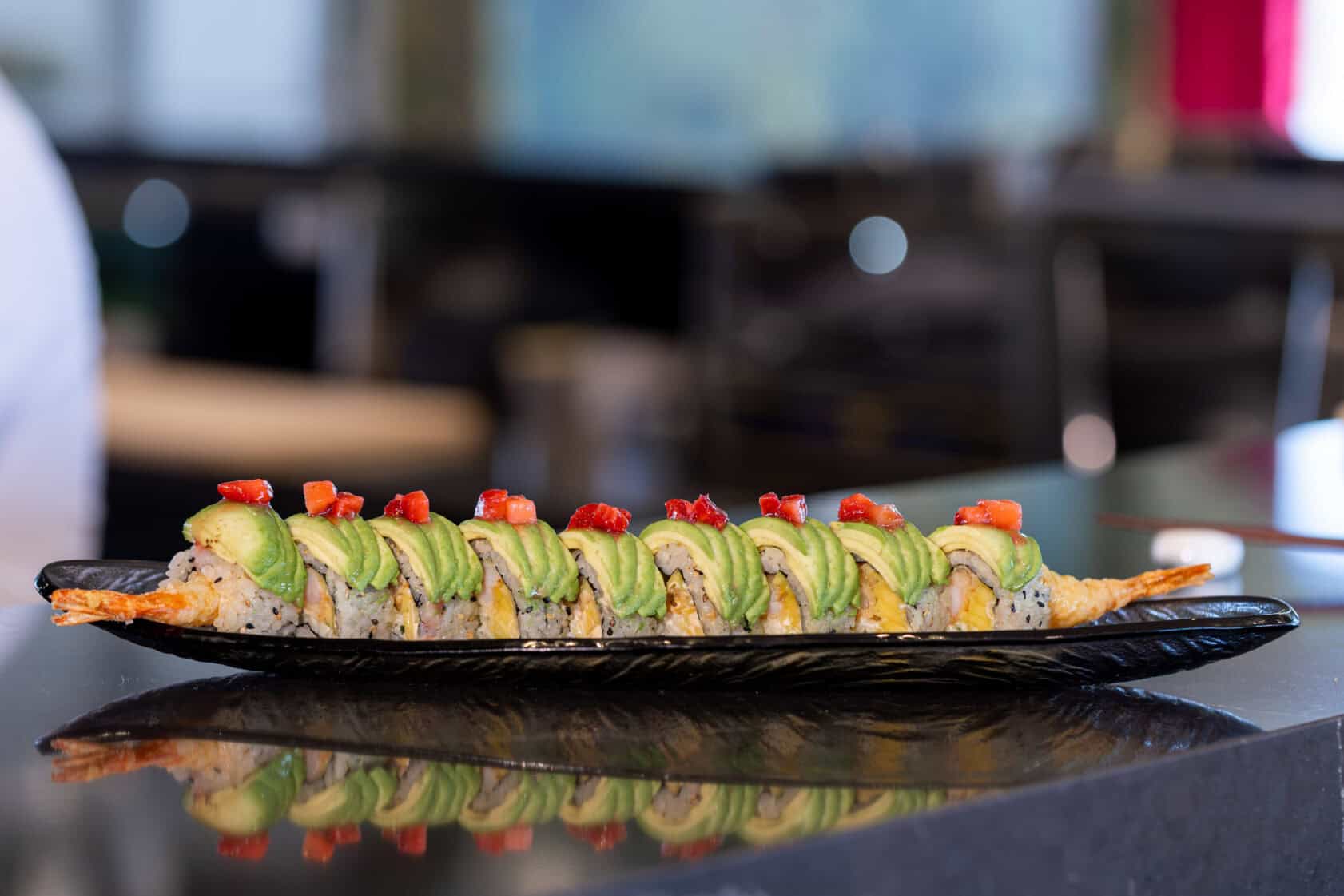 A sushi roll topped with sliced avocado and red garnish is served on a black rectangular plate, placed on a reflective countertop.