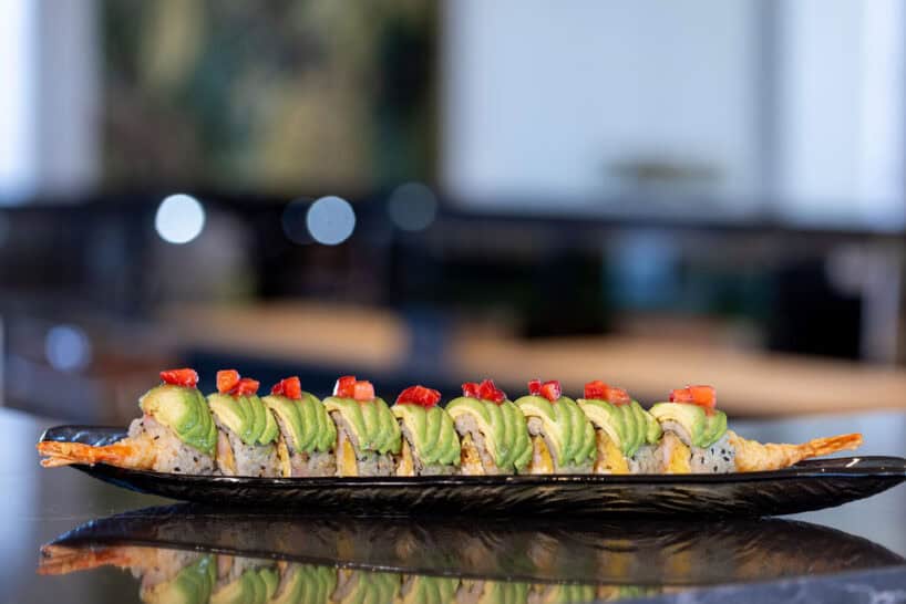 A sushi roll topped with sliced avocado and diced red pepper is arranged on a black plate, displayed on a reflective surface.