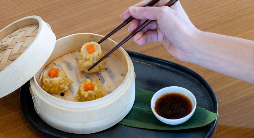 A hand uses chopsticks to pick up a shumai dumpling from a bamboo steamer basket, with a small dish of soy sauce on a black tray nearby.