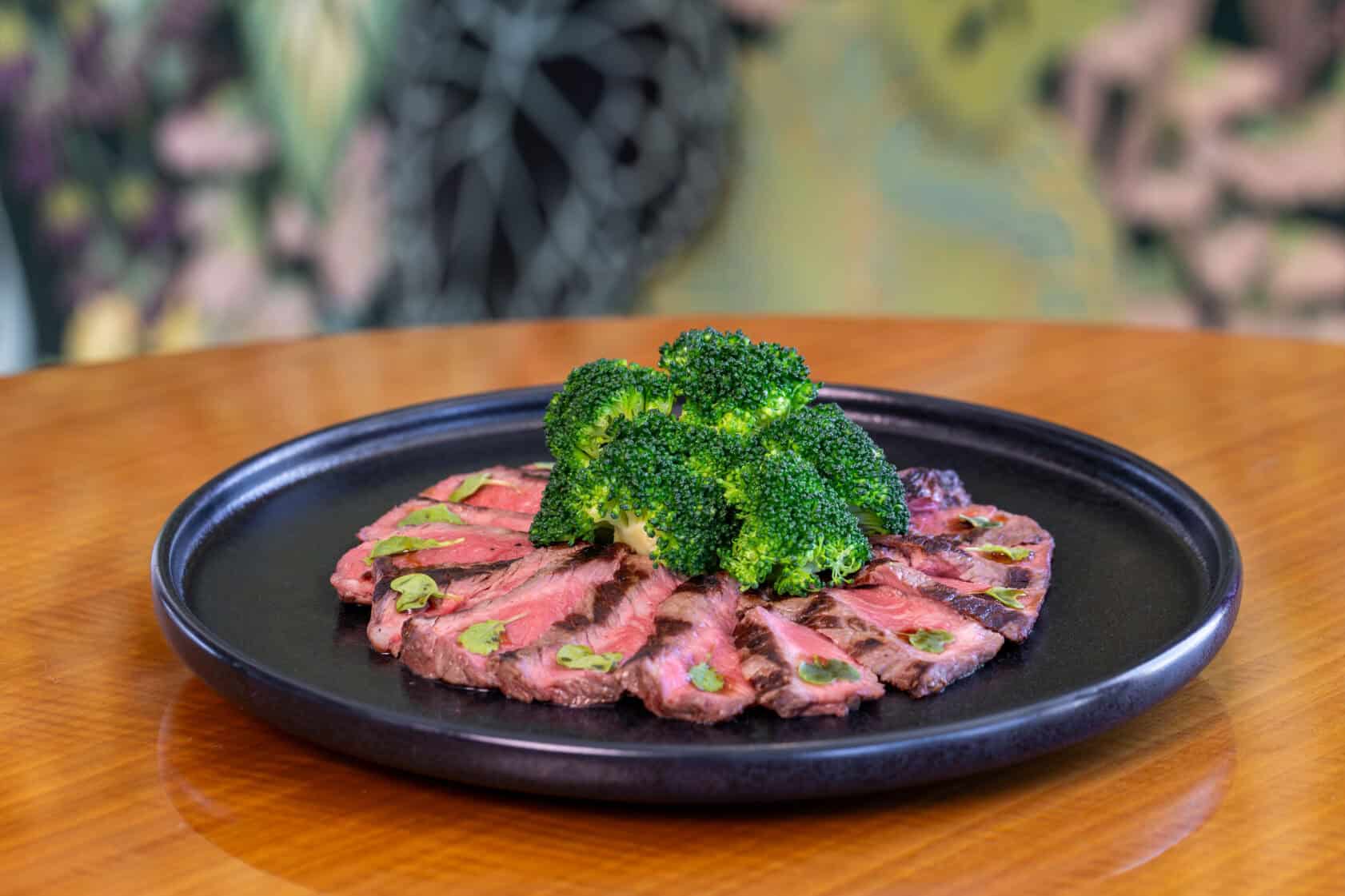 Sliced grilled steak arranged in a circle on a black plate, topped with steamed broccoli, set on a wooden table.