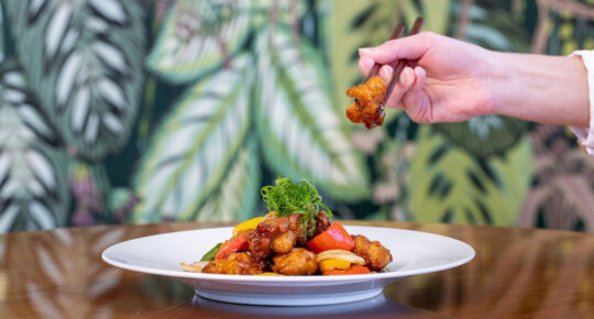 A hand uses chopsticks to pick up a piece of stir-fried chicken from a plate garnished with vegetables, set on a wooden table with a leafy patterned background.