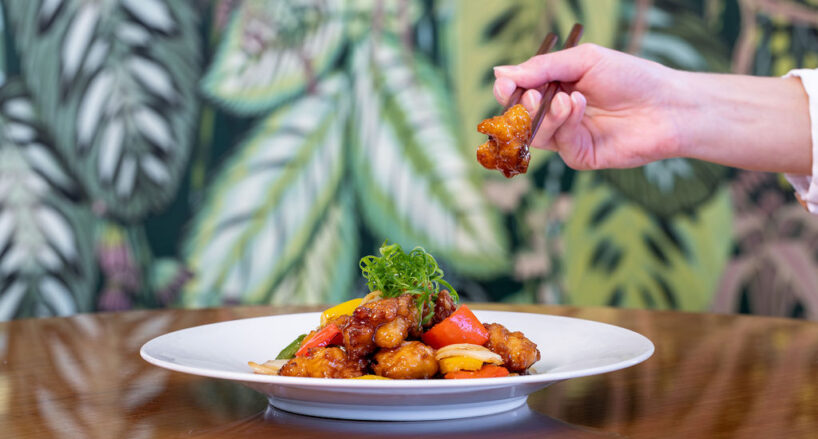 A hand uses chopsticks to pick up a piece of stir-fried chicken from a plate garnished with vegetables, set on a wooden table with a leafy patterned background.