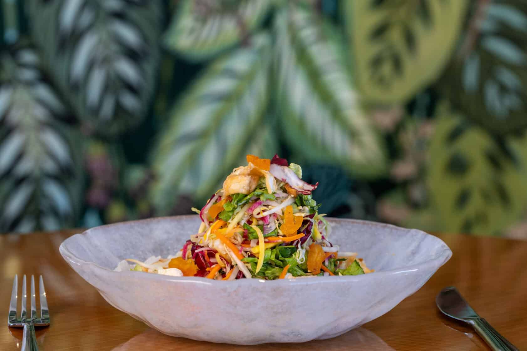 A colorful mixed salad with shredded vegetables and citrus slices is served in a shallow white bowl on a wooden table, with a fork and knife beside it.