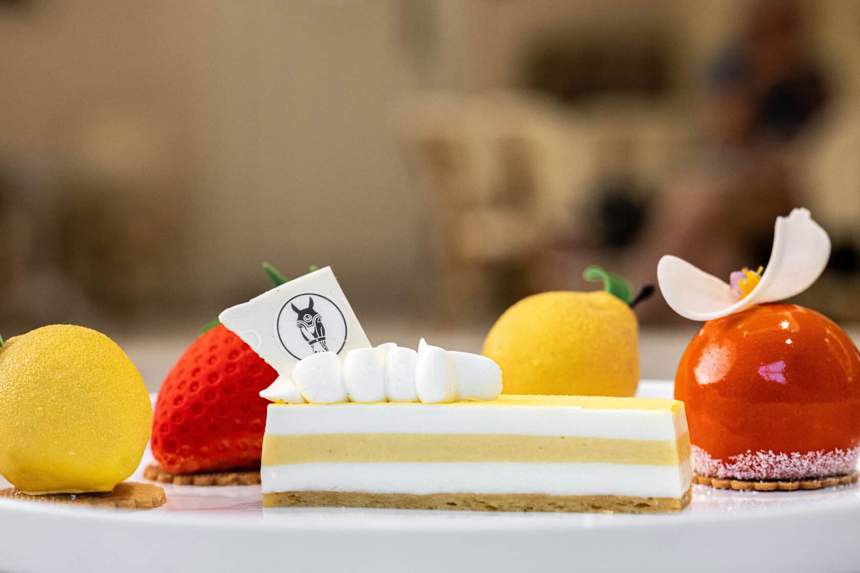 A plate with four colorful pastries, including a layered rectangular cake with white icing, and three dome-shaped desserts resembling fruit, set against a blurred background.