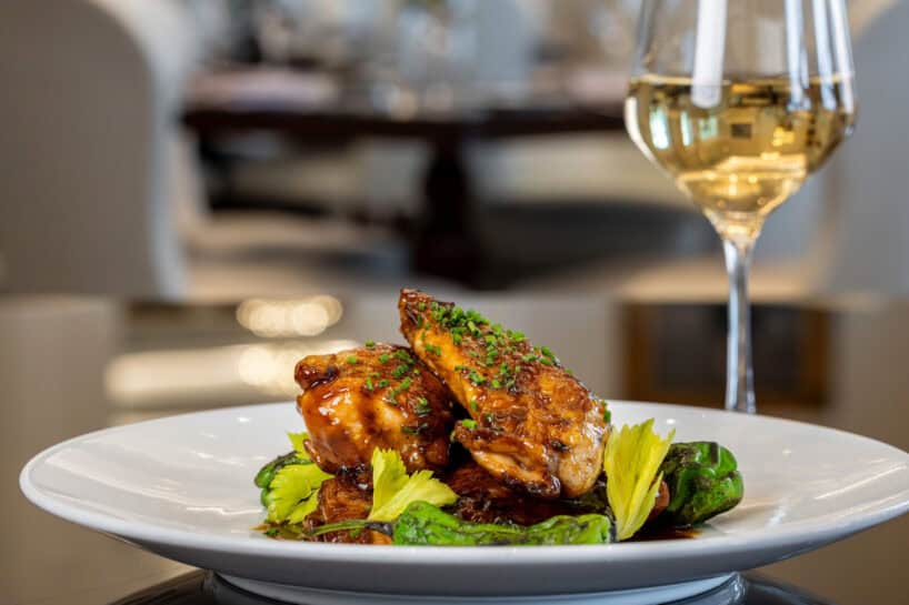 A plated dish of glazed chicken breast on vegetables, garnished with herbs, sits next to a glass of white wine in a restaurant setting.