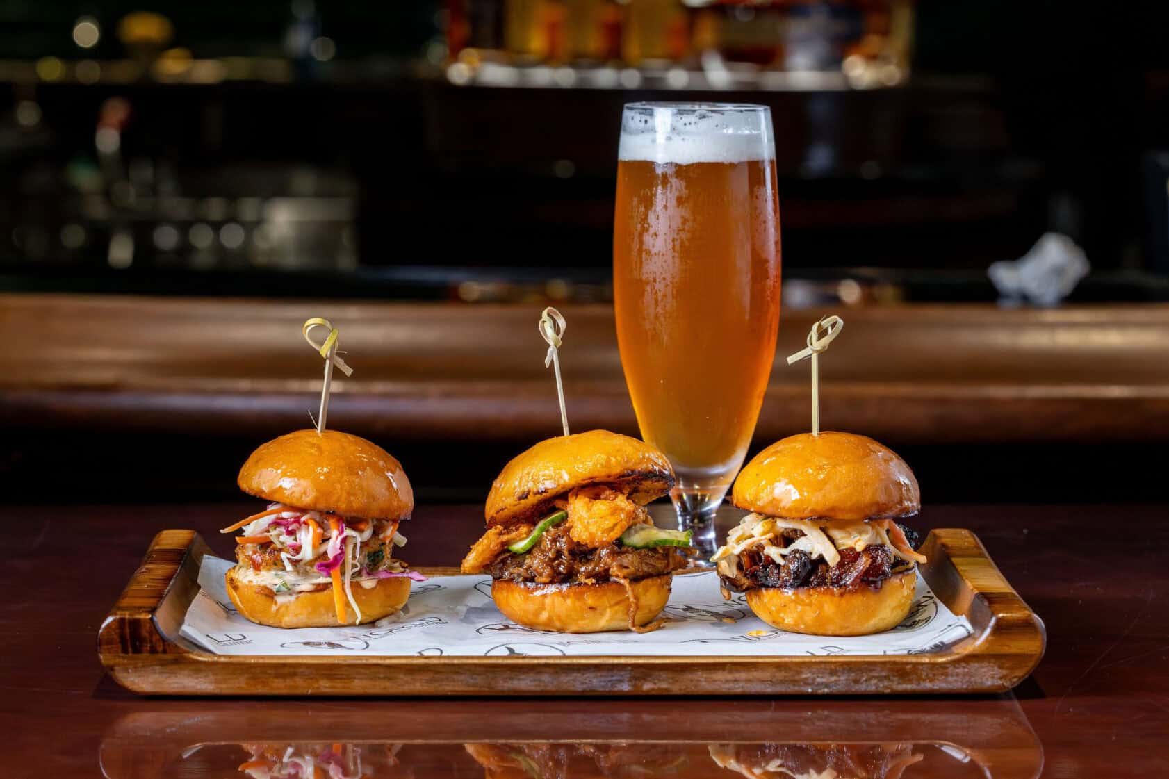 Three gourmet sliders with various toppings are arranged on a tray, each secured with a skewer, with a glass of beer in the background.