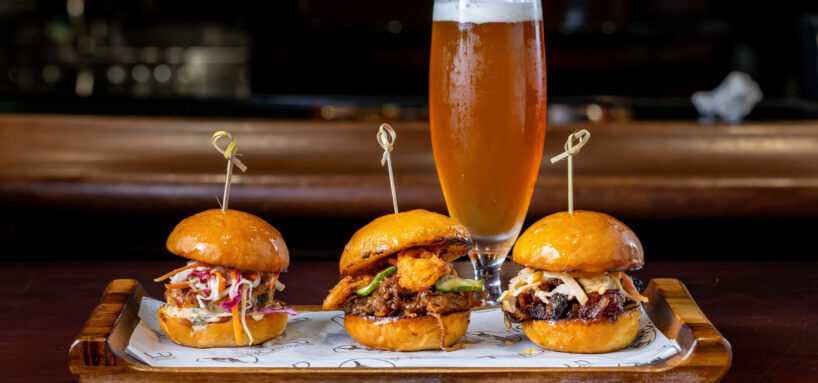 Three gourmet sliders with various toppings are arranged on a tray, each secured with a skewer, with a glass of beer in the background.