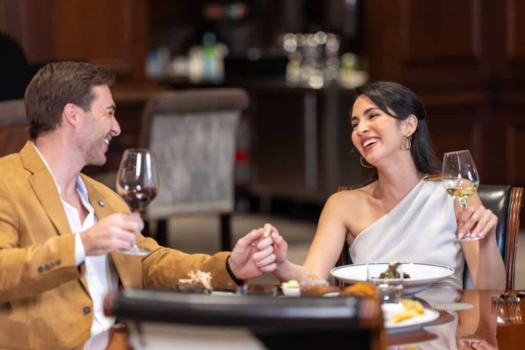 A man and woman sit at a restaurant table, holding hands, smiling, and toasting with wine glasses.