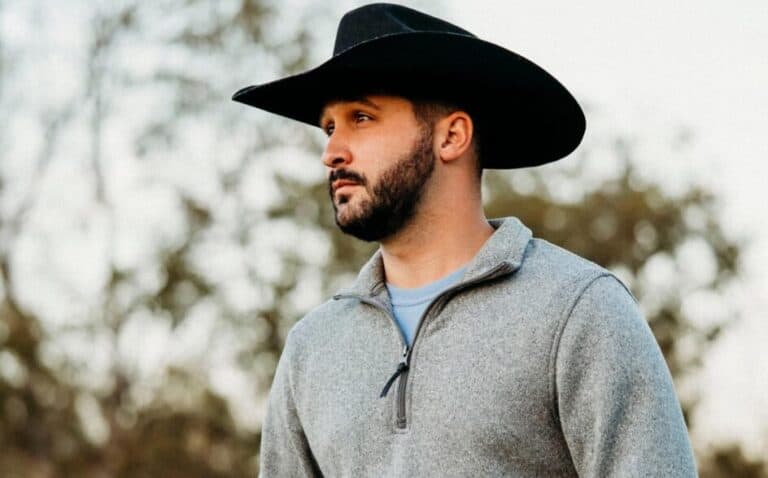 A man with a beard wearing a black cowboy hat and a gray zip-up sweater stands outdoors with trees in the background.