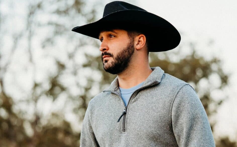 A man with a beard wearing a black cowboy hat and a gray zip-up sweater stands outdoors with trees in the background.