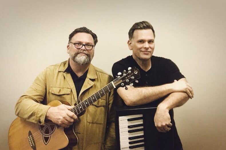 Two men posing indoors; one holds an acoustic guitar, the other leans on a keyboard. Neutral background.