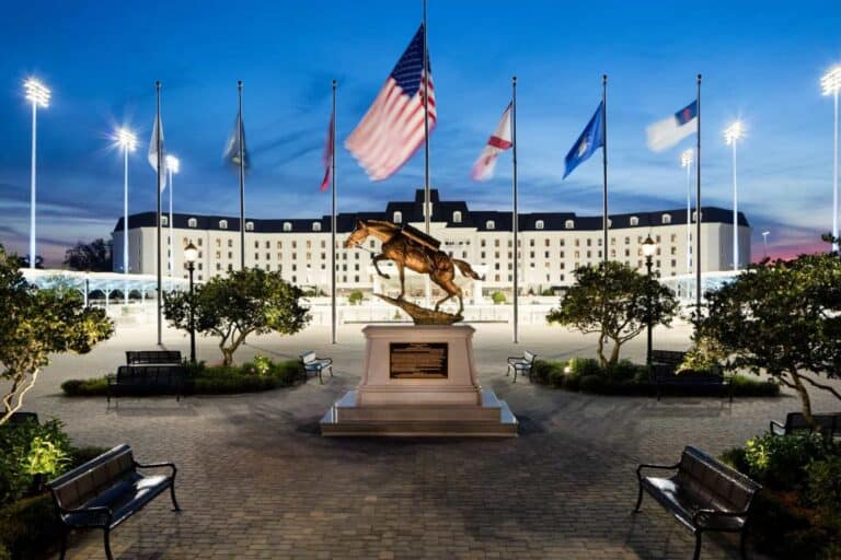 A bronze horse statue stands in front of a large hotel, surrounded by flags and benches, with bright lights illuminating the scene at dusk.
