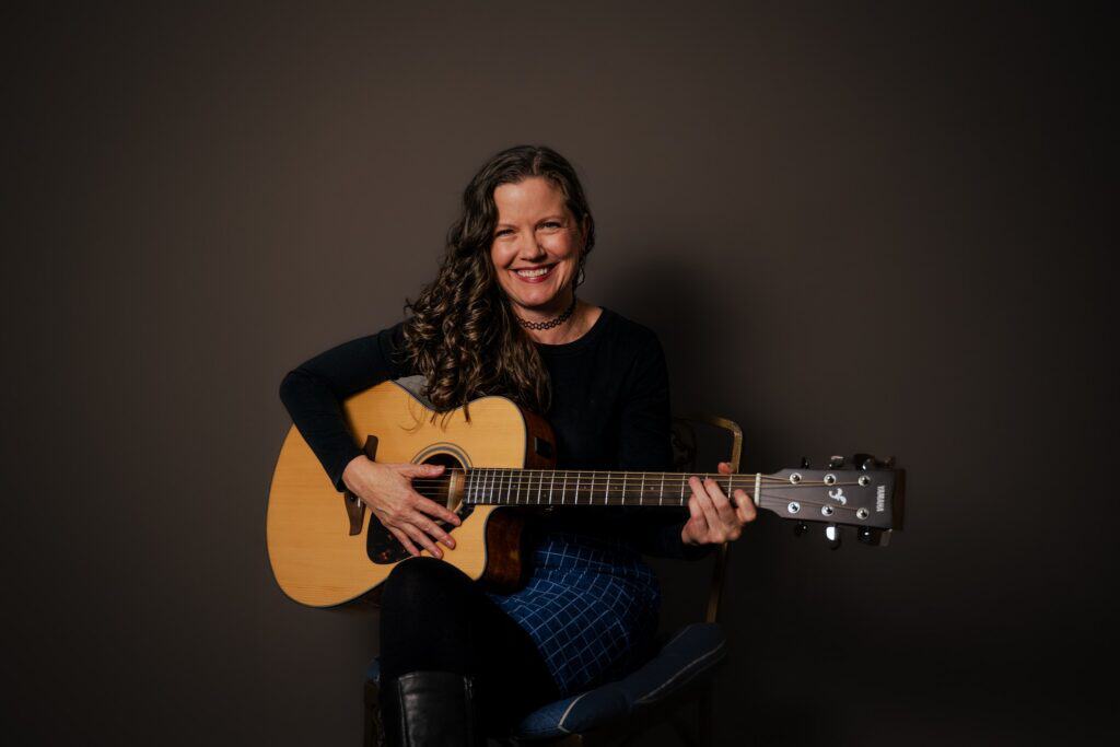 A woman with long, wavy hair sits on a chair, smiling and playing an acoustic guitar against a plain dark background.