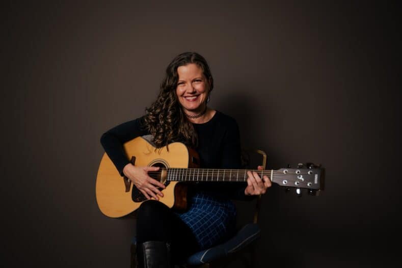 A woman with long, wavy hair sits on a chair, smiling and playing an acoustic guitar against a plain dark background.