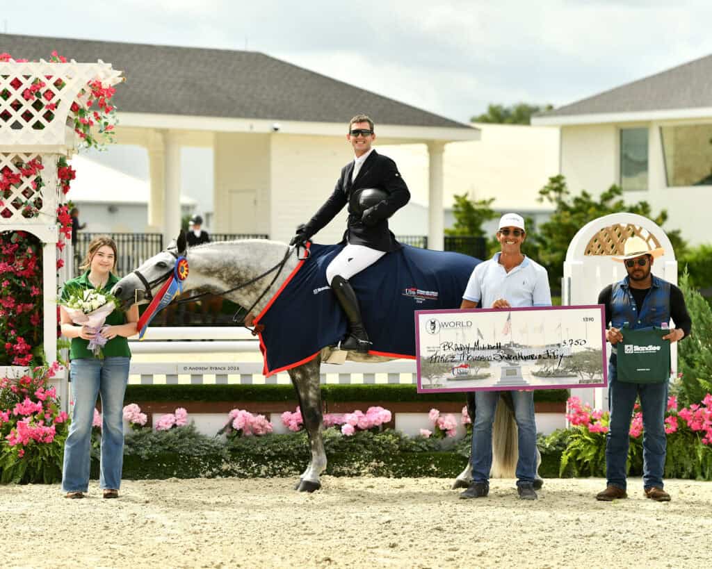 A man on horseback wears a winner's sash and is flanked by three people holding flowers, a large check, and a tote bag at an outdoor equestrian event.