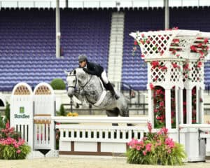 Equestrian rider on a gray horse jumps over a white and pink obstacle in an outdoor arena with empty blue stadium seats in the background.