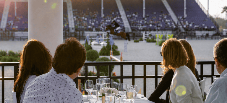Four people seated at an outdoor table watch an equestrian event, with a horse and rider jumping in a large arena filled with spectators.