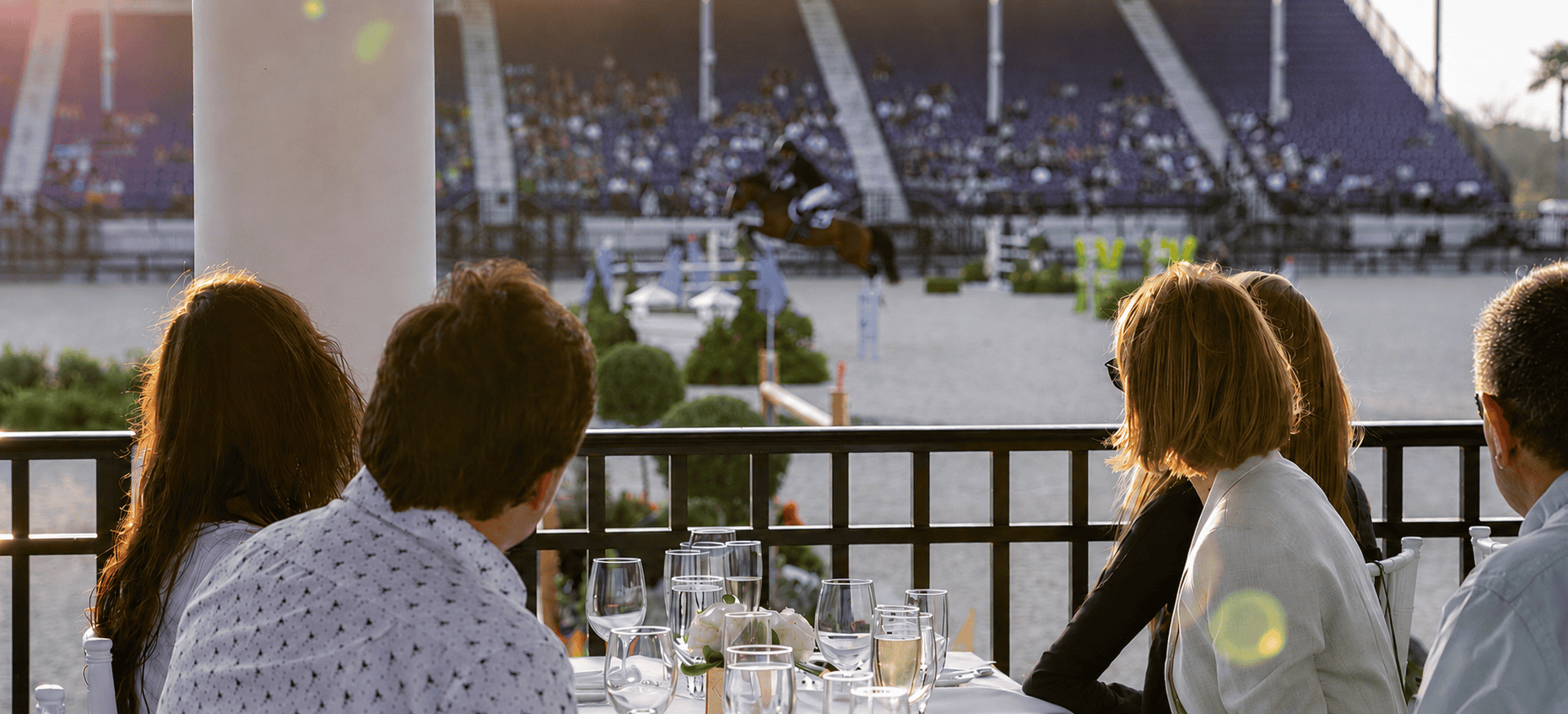 Four people seated at an outdoor table watch an equestrian event, with a horse and rider jumping in a large arena filled with spectators.