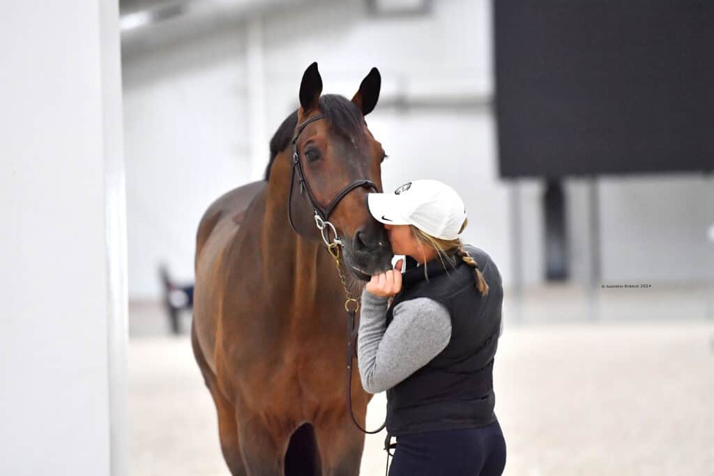 A person wearing a white cap and black vest stands close to a brown horse indoors, holding its bridle and touching its muzzle.
