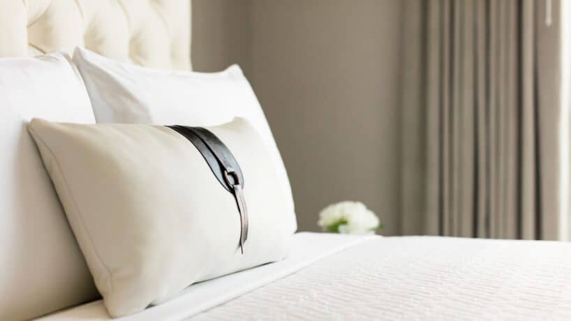 A neatly made bed with a white pillow featuring a black decorative strap, white bedding, and curtains in the background.