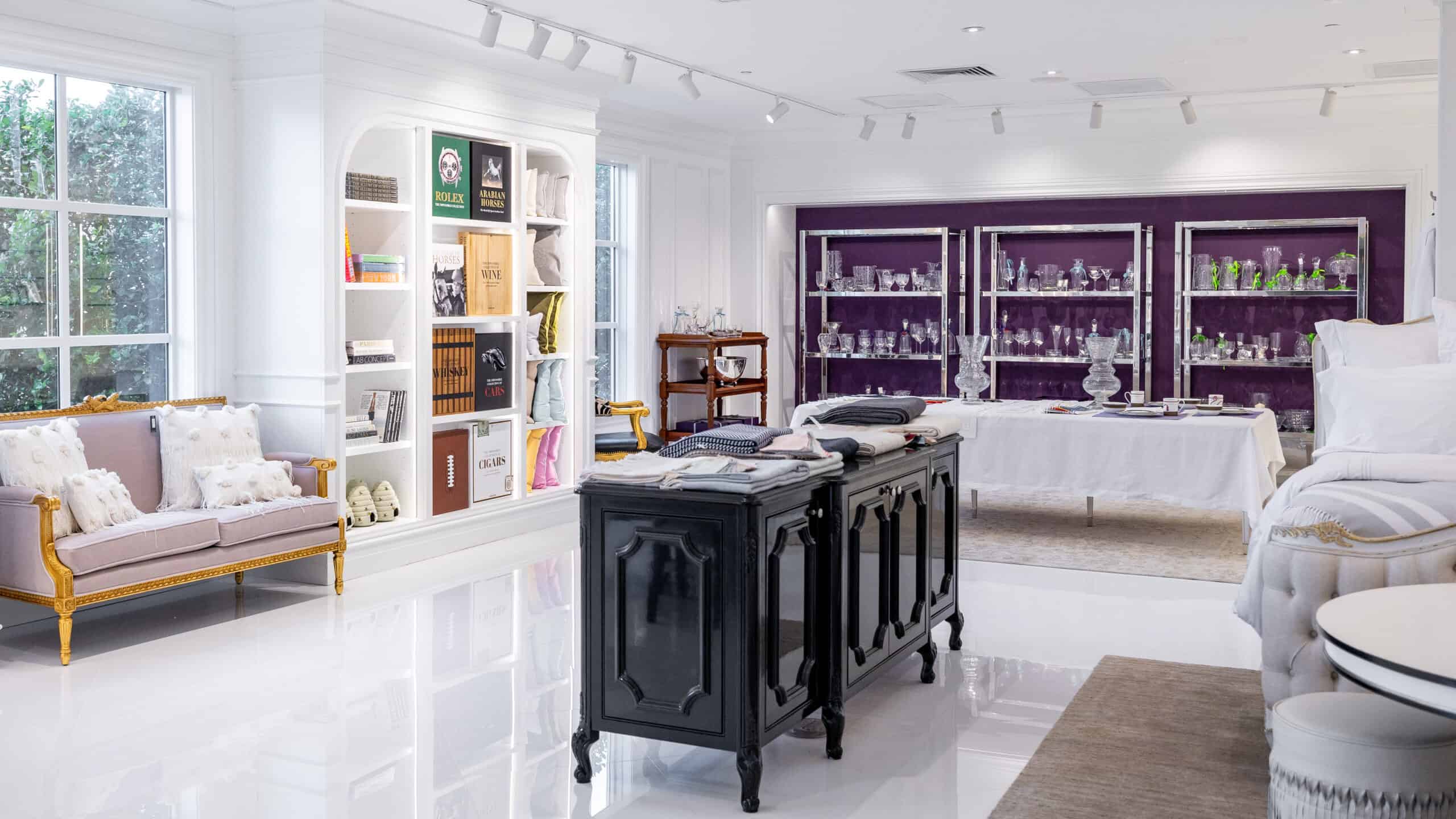 A bright, elegant showroom with white walls, a black island display, glass shelves of crystal items, bookshelves, and a gold-trimmed sofa near large windows.