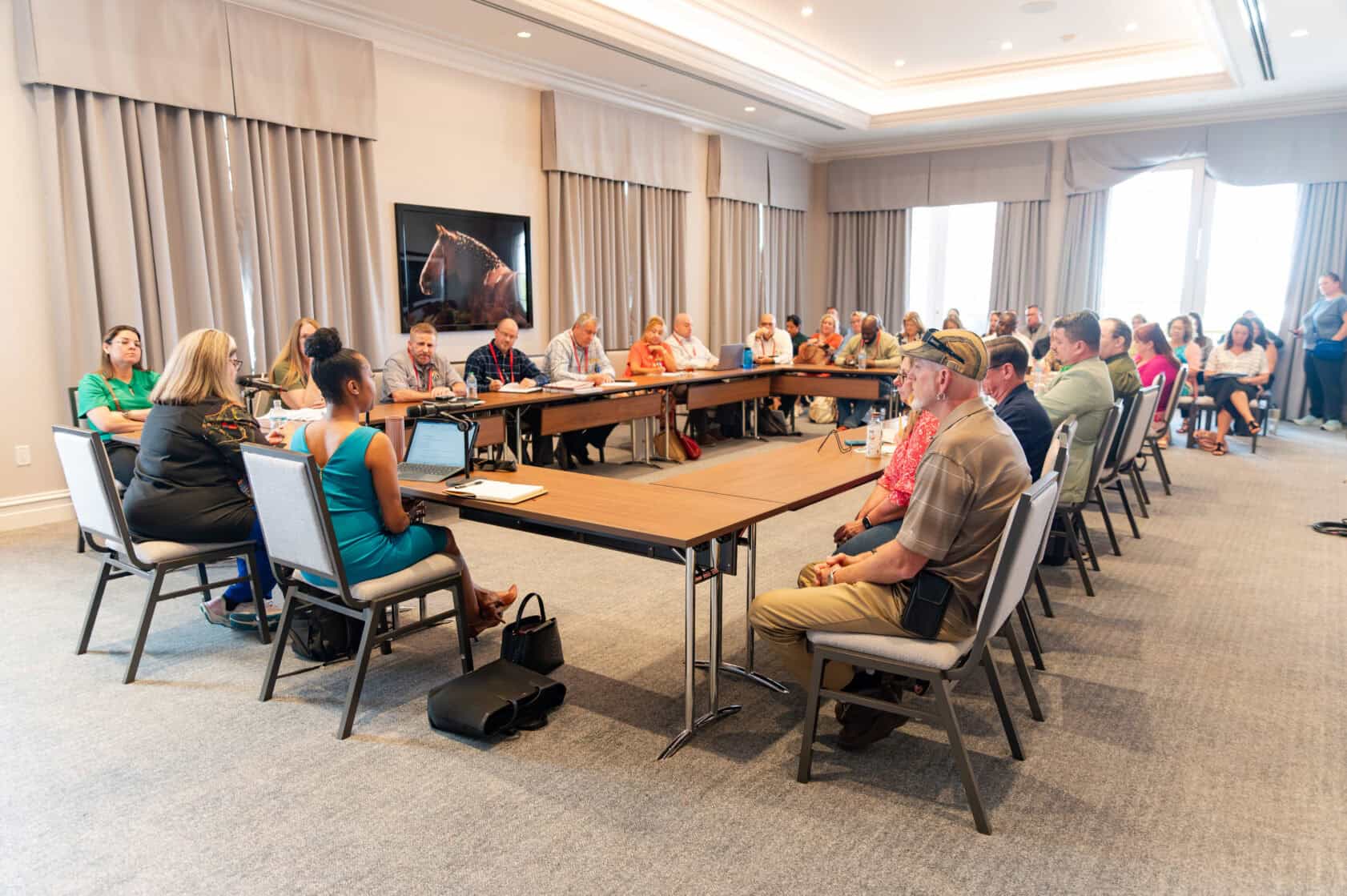 A group of people sit around a U-shaped table in a conference room, engaged in a meeting or discussion.