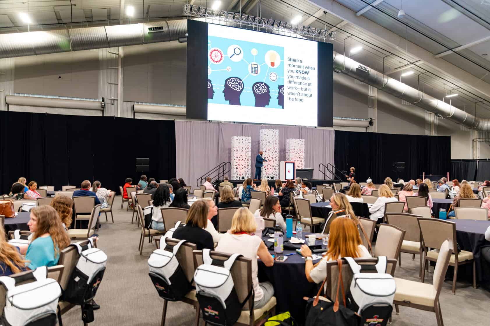 A speaker presents on stage to an audience seated at round tables in a large conference hall, with a slideshow displayed on a screen behind them.