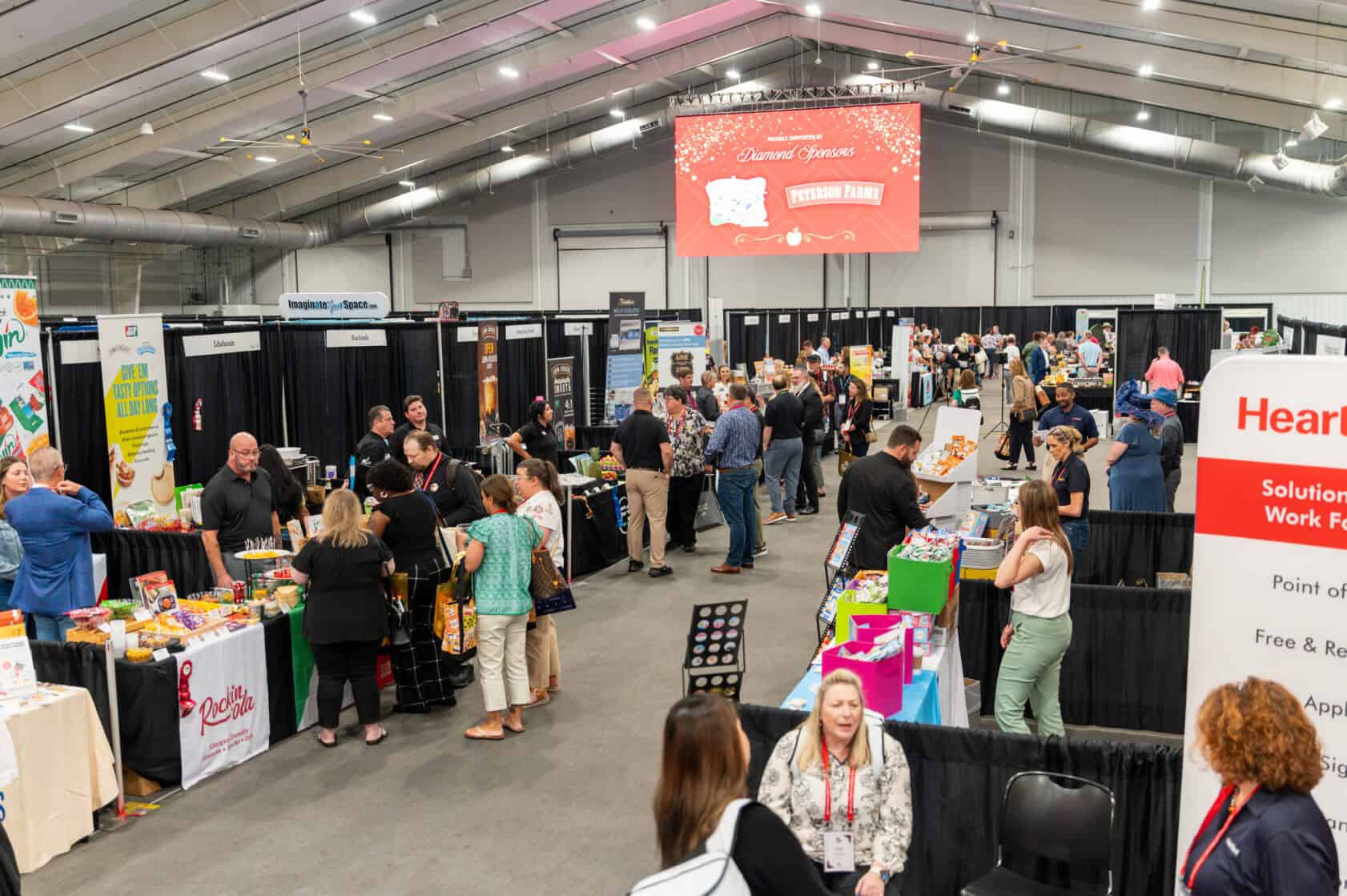 A crowded indoor convention with vendor booths, attendees walking and browsing, and a large red screen displaying event information at the back of the hall.