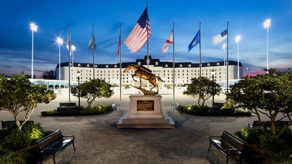 A horse statue stands in front of a large, well-lit building at dusk, with multiple flags on poles and benches lining a paved walkway.