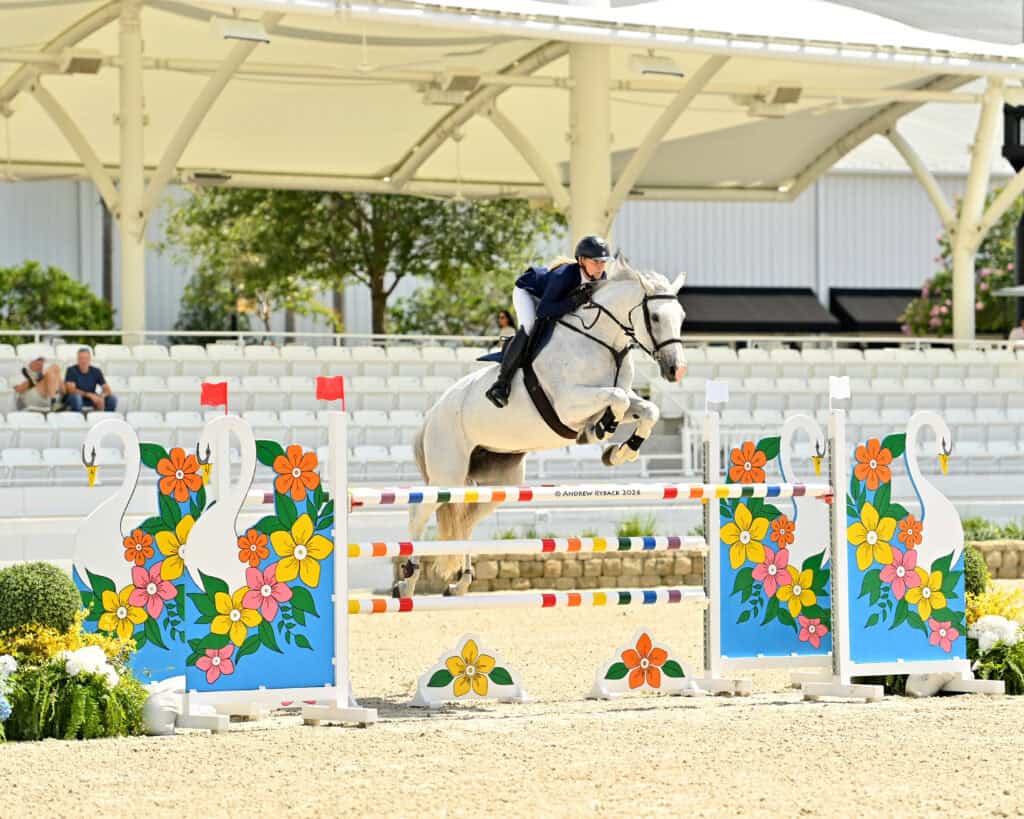 A rider on a white horse jumps over a colorful floral obstacle during an equestrian show jumping competition in an outdoor arena.