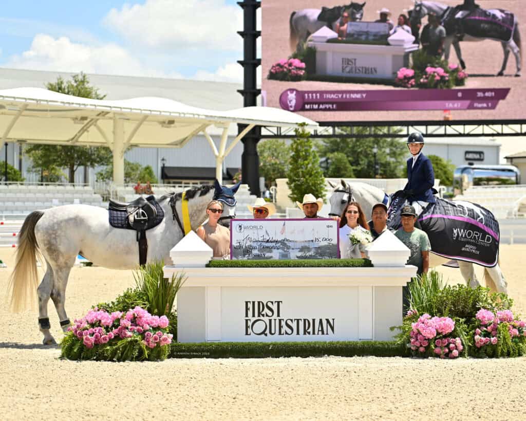 A group of people and two horses stand around a podium with "First Equestrian" written on it at an outdoor event, with flowers and a large event screen in the background.