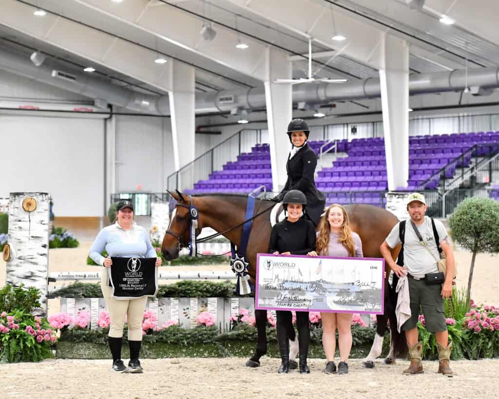 Five people and a horse pose indoors at an equestrian event; one person on horseback holds ribbons, others hold a trophy bag and an oversized check.