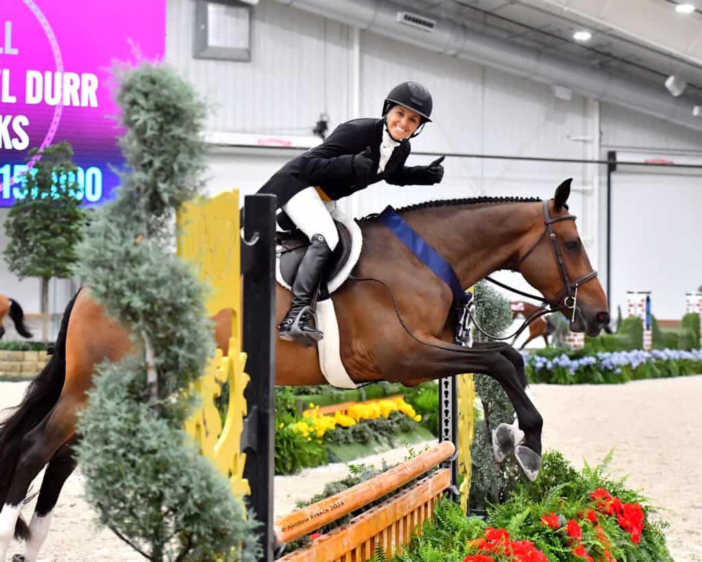 A rider in equestrian gear gives a thumbs up while jumping a horse over an obstacle during an indoor show jumping competition.