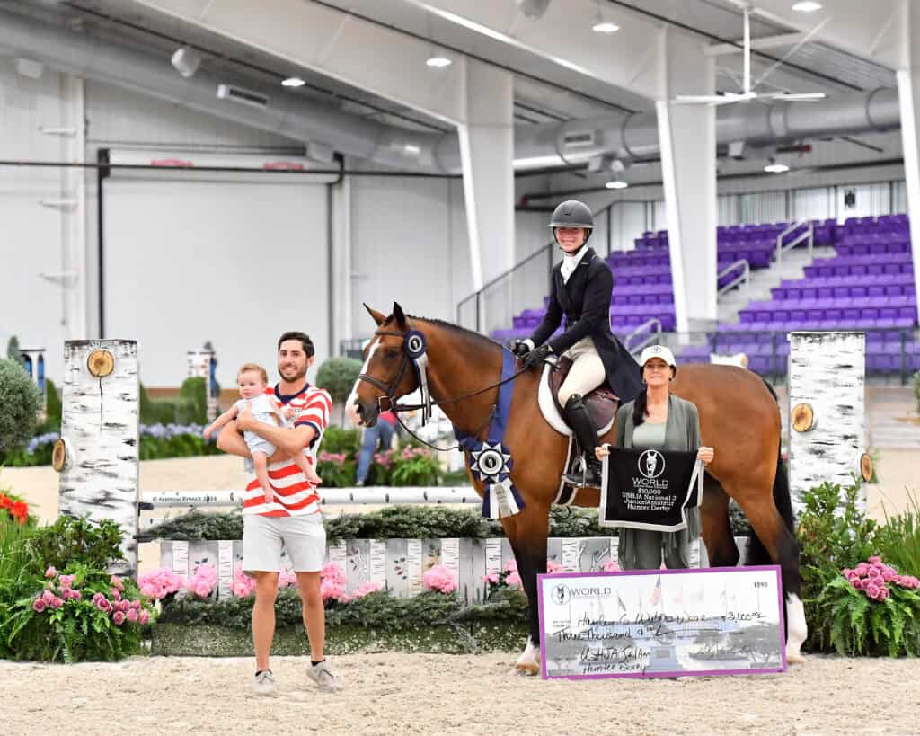 A woman on horseback poses with a ribbon beside a woman holding a large check, while a man holding a child stands nearby in an indoor arena with jumps and flowers.