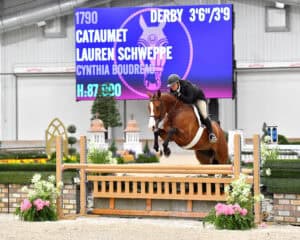 A rider in equestrian attire jumps a brown horse over a wooden obstacle in an indoor arena with a scoreboard in the background.