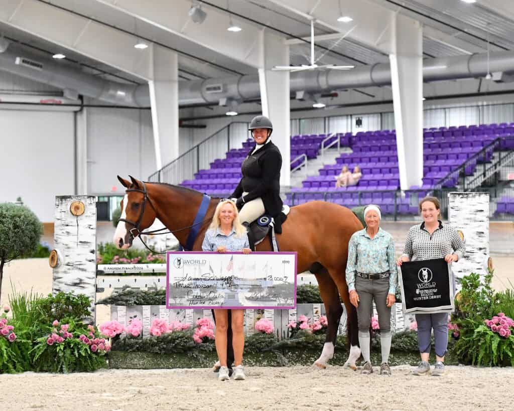 Four women and a horse pose indoors; one woman is mounted, another holds a large check, and others stand beside a banner. There are flowers and stadium seating in the background.