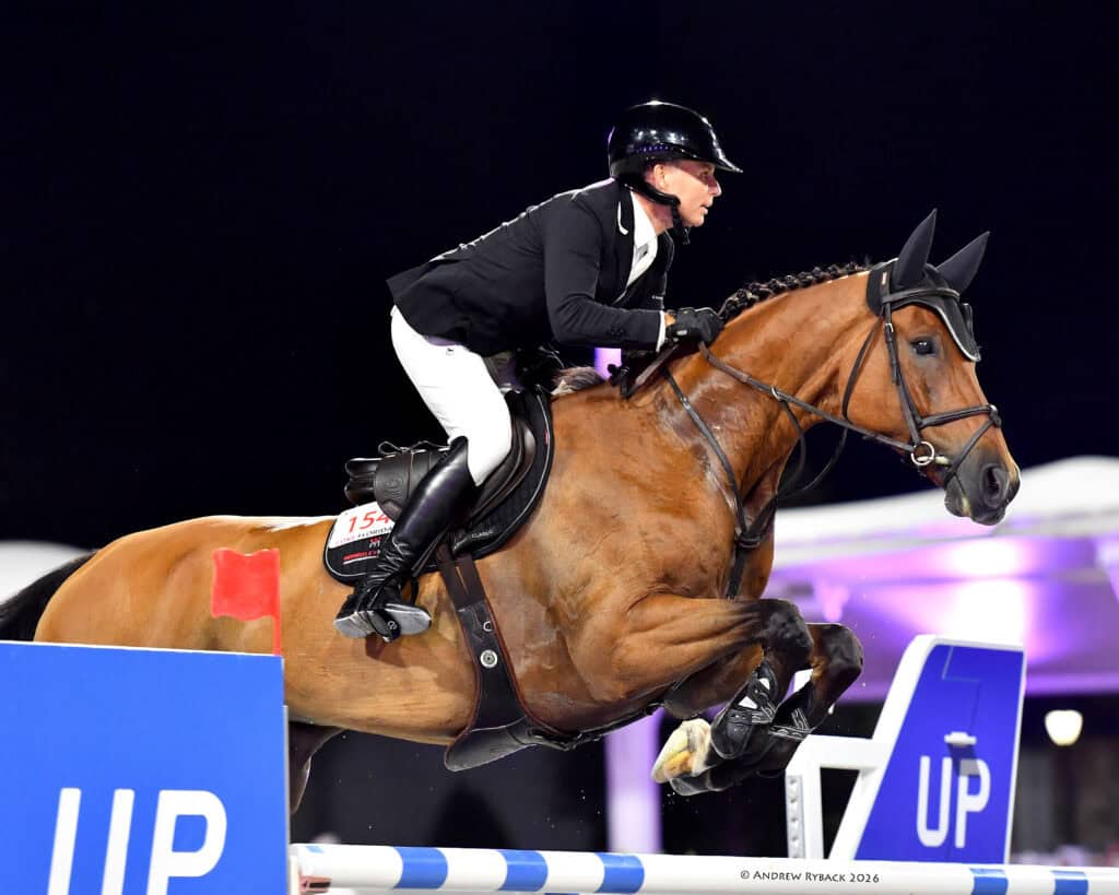 Equestrian rider in black jacket and helmet jumps a brown horse over a blue and white obstacle during a show jumping competition.