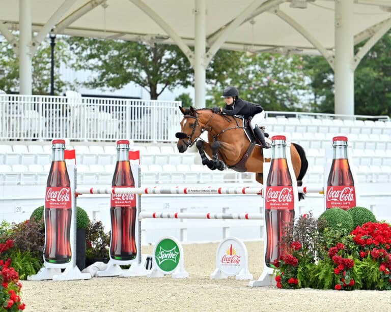 A rider on a horse jumps over an obstacle decorated with Coca-Cola and Sprite branding in an empty equestrian arena.