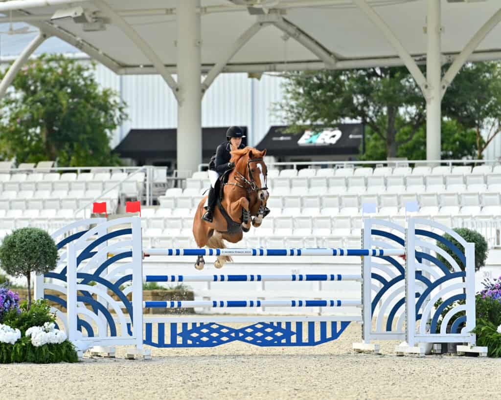 A rider in equestrian attire guides a horse over a blue and white jump in an outdoor arena with empty grandstand seating in the background.