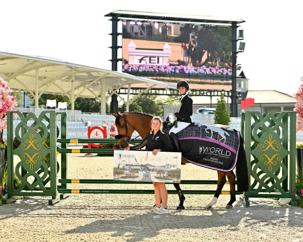 A rider on a horse poses with a rosette and a winner's blanket while another person holds a large check for 1,500 euros at an equestrian competition.