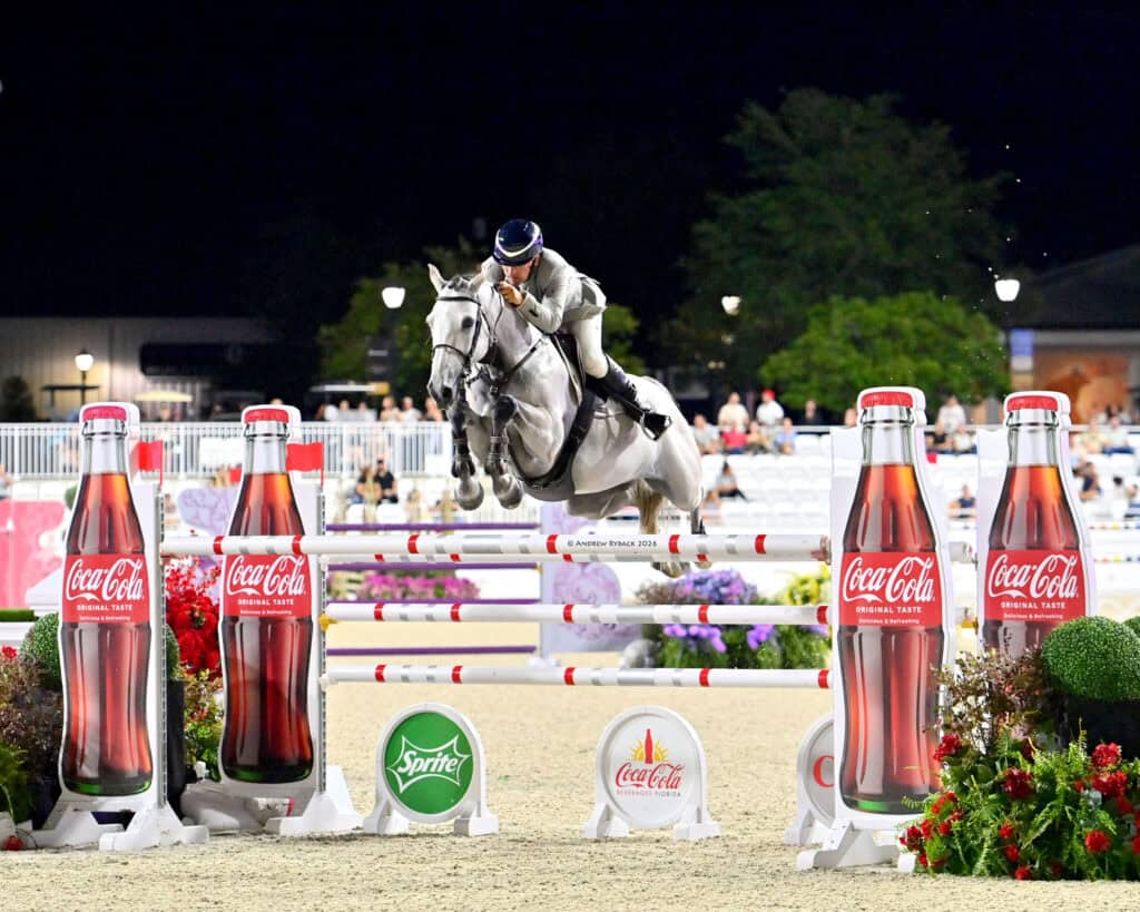 Equestrian rider on a gray horse jumps over an obstacle during a competition, with Coca-Cola and Sprite advertisements in the foreground.