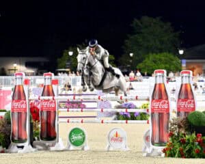 Equestrian rider on a gray horse jumps over an obstacle during a competition, with Coca-Cola and Sprite advertisements in the foreground.
