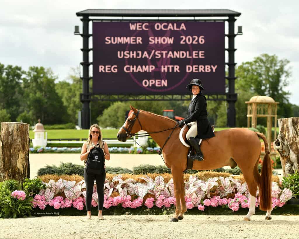 A rider on horseback and a woman standing beside them pose in front of a display board reading "WEC Ocala Summer Show 2026" at an outdoor equestrian event.