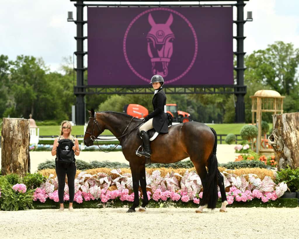 Equestrian rider on a horse poses for a photo with a handler at an outdoor event, with a large screen and floral decorations in the background.